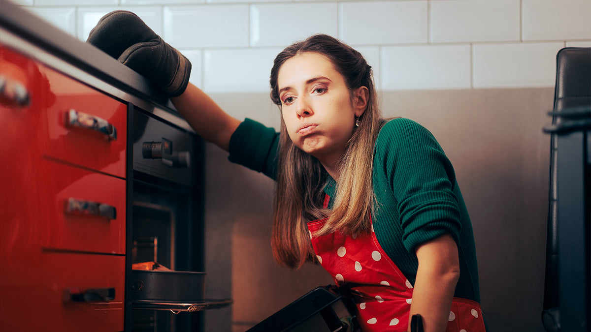 Woman in a kitchen wearing an apron and oven mitt, leaning against a red oven.