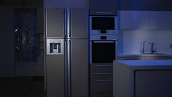 A dark kitchen with a faint blue light illuminating a silver fridge, black oven, and gray countertops.