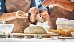 A couple wearing aprons sprinkling flour atop bread dough.