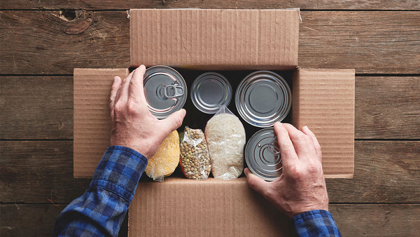 The hands of a man packing tin cans and bagged foods into a cardboard box placed on a wood table.